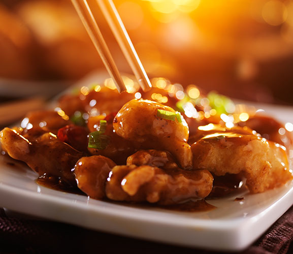 A close-up of a plate of delicious, saucy Asian chicken dish with chopsticks, illuminated by warm, golden light.