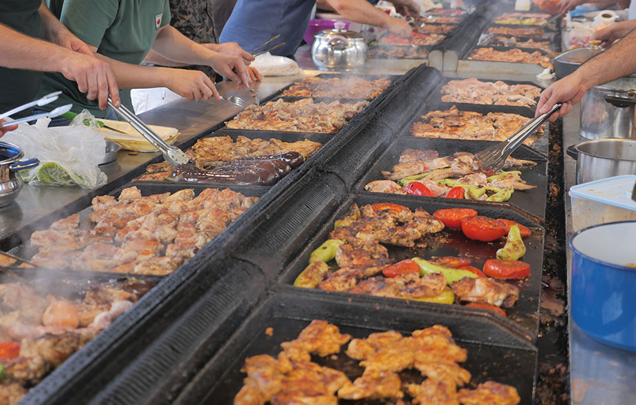 A bustling outdoor grill with chefs cooking various meats and vegetables, surrounded by steam and vibrant colors.