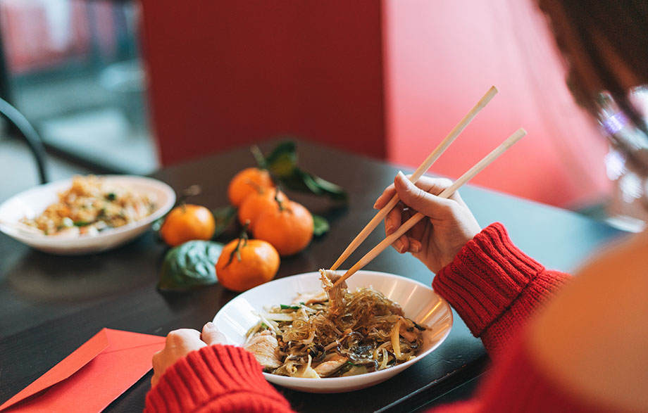 A person wearing a red sweater uses chopsticks to eat from a bowl of noodles, with tangerines and another dish in the background.