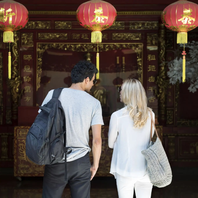 A couple stands in front of a richly decorated temple entrance, adorned with red lanterns, while gazing at the interior.