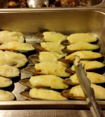 Baked mussels topped with melted cheese, arranged in a metal serving tray with tongs resting nearby.