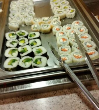 A metal tray displays assorted sushi rolls, including colorful vegetable and seafood variations, with serving tongs on the side.