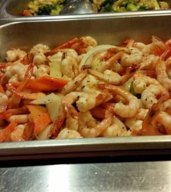 A metal tray filled with sautéed shrimp, mixed with colorful vegetables such as carrots and bell peppers, garnished with herbs.