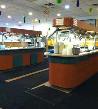 A spacious buffet restaurant interior featuring two serving stations, stacked plates, and festive decorations hanging from the ceiling.