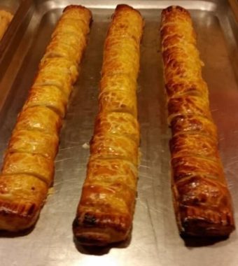 Three golden-brown pastry rolls topped with melted cheese, placed in a metal serving tray with onion rings in the background.