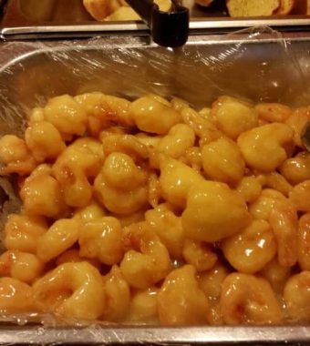 A serving tray filled with glossy, sweet shrimp in a light sauce, accompanied by a spoon for serving. Plastic wrap covers the dish.