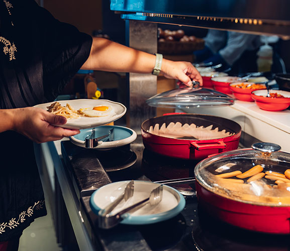 A person in a black shirt serves food from a buffet, holding a plate with an egg and noodles, while lifting a pot lid.