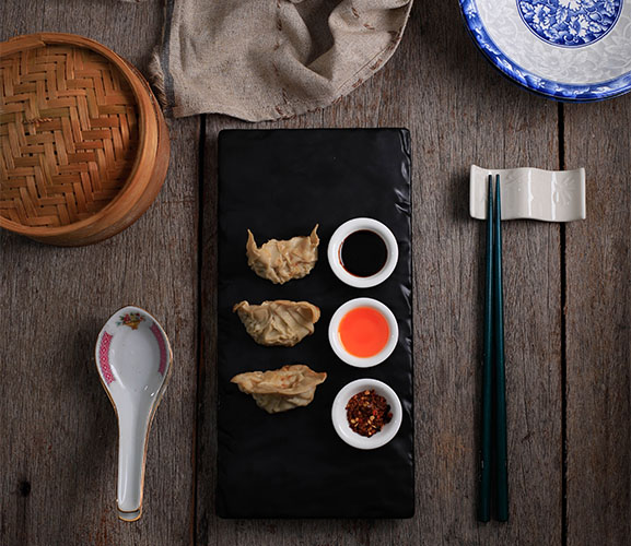 A black plate with steamed dumplings and three dipping sauces, surrounded by chopsticks, a spoon, and a bamboo steamer.