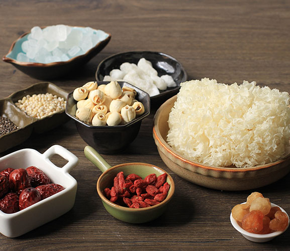 An array of traditional ingredients: dried fruits, lotus seeds, goji berries, and snow fungus displayed in various dishes on a wooden surface.